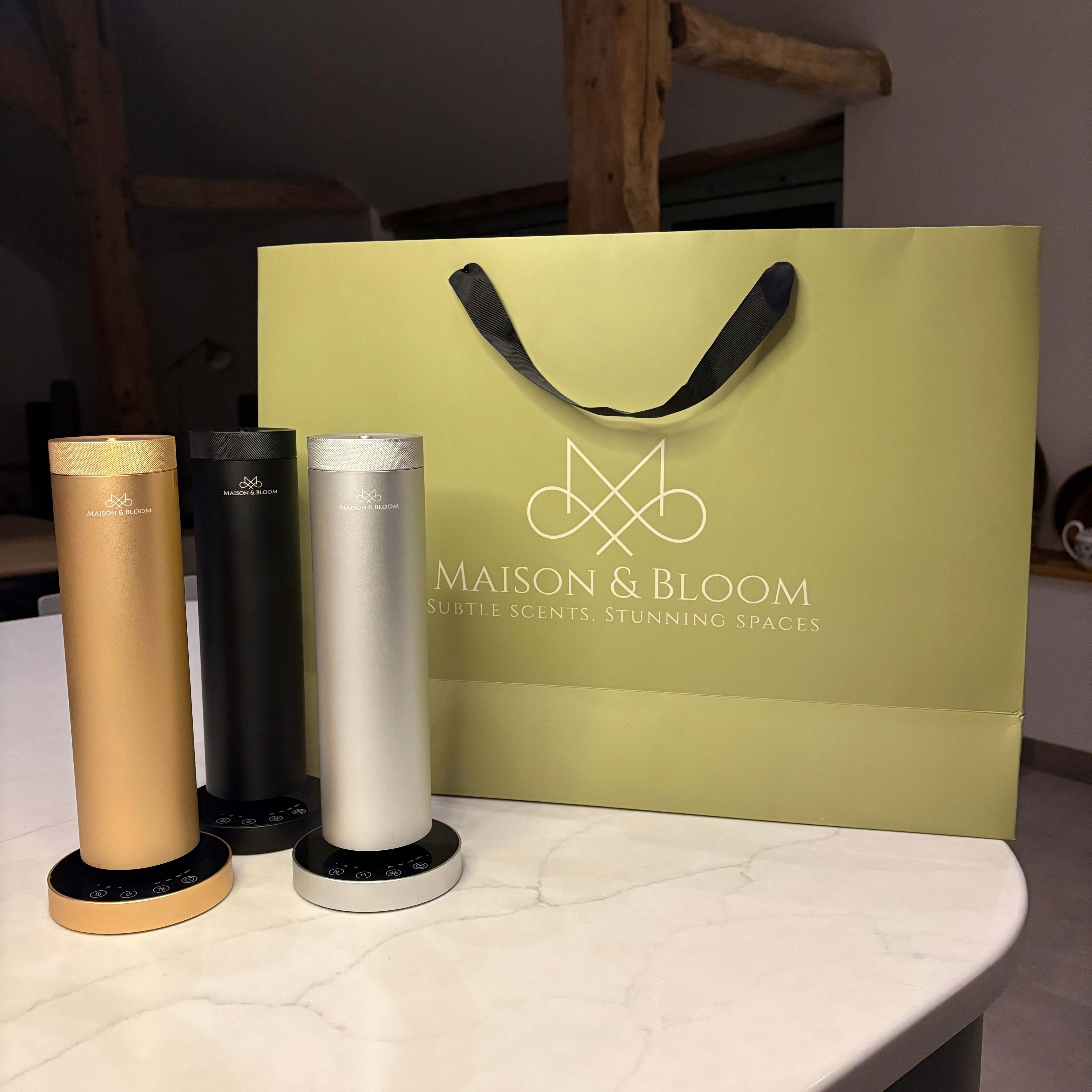 Three modern scent diffusers in gold, black, and silver on a counter with a gift bag.